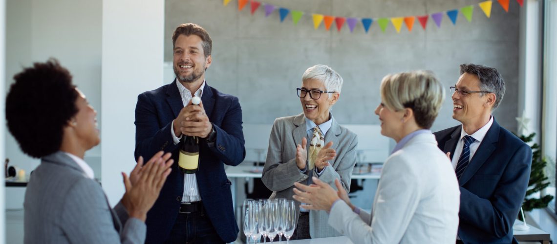 Group of happy colleagues celebrating business success and applauding while their colleague is opening bottle of Champagne in the office.